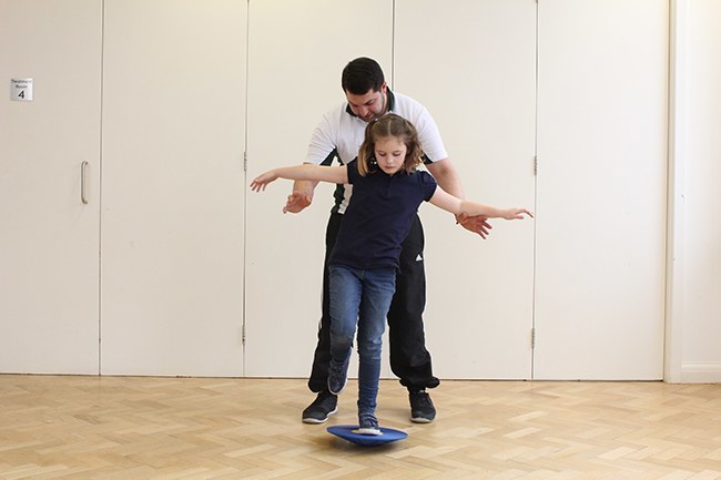 Child balancing on wobble board on one leg, Therapist behind for support