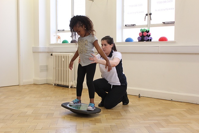 Child balancing on wobble board, therapist offering support